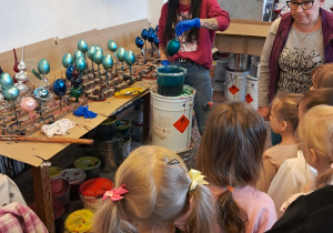 Grupa dzieci znajduje się w fabryce bombek. Obserwują one jak Pani farbuje bombki.