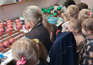 Grupa dzieci znajduje się w fabryce bombek. Obserują one jak dwie Panie ręcznie dekorują bombki.