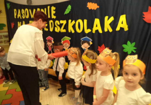 Na zdjęciu widoczne są dzieci oraz Pani Dyrektor, która dokonują pasowania Przedszkolaków, przy pomocy ogromnego ołówka.