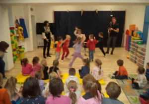 Na zdjęciu widać dzieci z naszego przedszkola oraz przybyłych Gości. Sześcioro dzieci tańczy krakowiaka do granej muzyki przez Pana Szymona. Pozostałe dzieci oglądają taniec kolegów z ogromnym zaciekawieniem