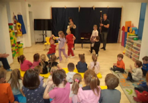 Na zdjęciu widać Panią i Pana Szymona, który gra krakowiaka na fagocie. Są również dzieci, które tańczą krakowiaka oraz pozostałe przedszkolaki, które oglądają występ swoich kolegów.