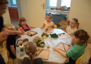 Grupa dzieci siedzi przy stole i przykleja gałązki do kwadratowych ramek.