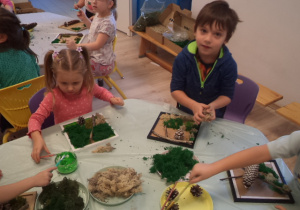 Dzieci siedzą przy stole i przyklejają mech do kwadratowych ramek.