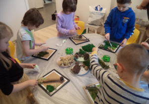 Dzieci stoją przy stoliku i dekorują kwadratowe ramki.