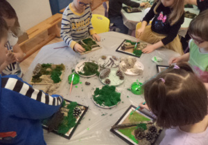 Dzieci stoją przy stoliki i dekorują kwadratowe ramki mchem.