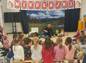 Dzieci siedzą w sali gimnastycznej. Obserwują pana siedzącego na tle dużego plakatu przedstawiającego łąkę. Nad dziećmi wisi napis Mikołajki.