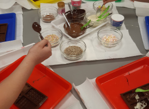 Na zdjęciu widoczny jest stolik z roztopionymi czekoladami i dodatkami do ich ozdabiania.