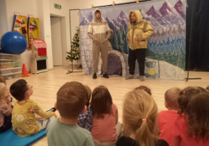 Dwóch aktorów ubranych w zimowe stroje stoi na tlezimowej scenografii.