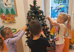 Dwie dziewczynki i chłopiec ubierają choinkę.