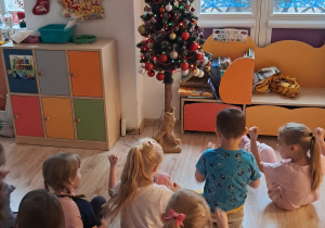 Dzieci siedzą na podłodze i podziwiają ubraną choinkę.