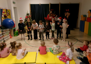 Na zdjęciu można zobaczyć dwie Panie prowadzące grudniową audycję muzyczną oraz dzieci, które śpiewają, naśladują pokazywane ruchy oraz dzwonią dzwoneczkami - niczym elfy Świętego Mikołaja.