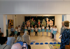 Na zdjęciu widoczne są dzieci oraz Panie Nauczycielki z grupy Motylków, którzy uczestniczą w świątecznym spotkaniu artystycznym. Dziewczynki ubrane są w niebieskie spódniczki i białe bluzki, a chłopcy w ciemne spodnie i białe koszule.