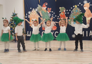 Na zdjęciu widoczne są dzieci oraz Panie Nauczycielki z grupy Motylków, którzy uczestniczą w świątecznym spotkaniu artystycznym. Dziewczynki ubrane są w zielone spódniczki i białe bluzki, a chłopcy w ciemne spodnie, białe koszule i zielone czapki choinki.