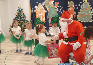Na zdjęciu widać Mikołaja, który wręcza dziewczynce prezent. Pozostałe dzieci obserwują Mikołaja.