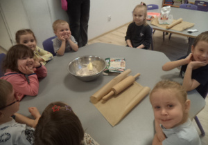 Na zdjęciu widać dzieci, które siedzą przy stole. Przed nimi stoi miska ze składnikami na pierniczki, wałek i papier do pieczenia.