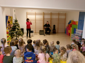 Na zdjęciu widać Panią prowadzącą audycję oraz Pana grającego na gitarze elektrycznej oraz obserwujące ich dzieci.