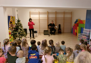 Na zdjęciu widać Panią prowadzącą audycję oraz Pana grającego na gitarze elektrycznej oraz obserwujące ich dzieci.