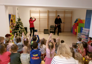 Na zdjęciu widać Panią prowadzącą audycję, Pana grającego na gitarze elektonicznej oraz dzieci naśladujące ruchy Pani.