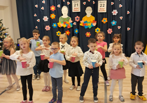 Na zdjęciu widać elegancko ubrane dzieci stojące w trzech rzędach. Każde dziecko trzyma kolorowy kubeczek.