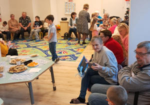 Na zdjęciu widać babcie, dziadków i dzieci siedzących w sali.