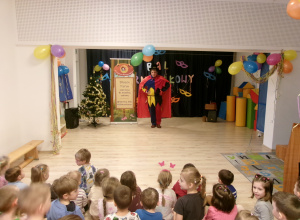 Na zdjęciu widać dzieci siedzące na sali, które obserwują kolorowo ubranego magika.