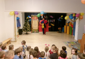 Na zdjęciu widać dzieci siedzące na sali, które obserwują kolorowo ubranego magika.