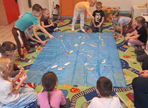 Na zdjęciu widać dzieci, które siedzą dokoła niebieskiego materiału. Dwójka dzieci łowi znajdujące się na materiale rybki.