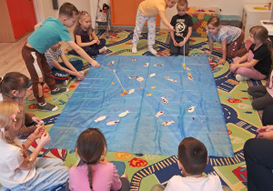 Na zdjęciu widać dzieci, które siedzą dokoła niebieskiego materiału. Chłopiec i dziewczynka łowią znajdujące się na materiale rybki.