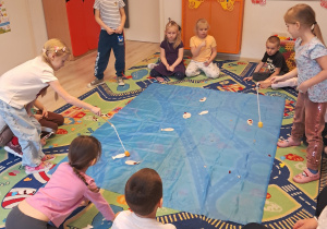 Na zdjęciu widać dzieci, które siedzą dokoła niebieskiego materiału. Dwie dziewczynki łowią znajdujące się na materiale rybki.