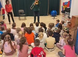 Na zdjęciu widać dzieci, które siedzą na sali gimnastycznj i słuchają Pana, który gra na puzonie.