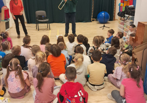 Na zdjęciu widać dzieci, które siedzą na sali gimnastycznj i słuchają Pana, który gra na puzonie.