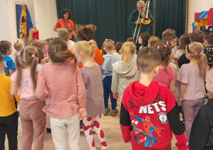 Na zdjęciu widać dzieci, które tańczą do akompaniamentu puzonu.