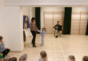 Na zdjęciu jest Pani prowadząca warsztaty wraz z Panem grającym na akordeonie i dziewczynką z grupy II. Dziewczynka z Panią tańczą.