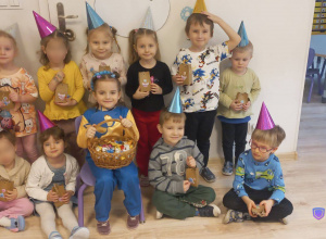 Na zdjęciu widać dzieci ubrane w kolorowe urodzonowe czapeczki, trzymające małe prezenciki. Na środku siedzi dziewczynka trzymająca koszyk z cukierkami.