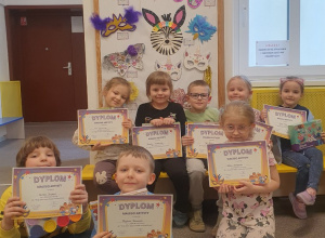 Na zdjęciu widać przedszkolaki, które pozują z dyplomami przy wystawie konkursowych masek.