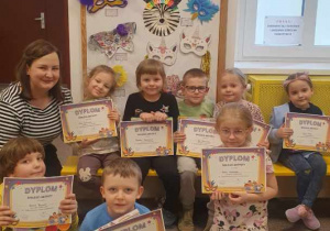 Na zdjęciu widać panią Monikę oraz przedszkolaki, które pozują z dyplomami przy wystawie konkursowych masek.