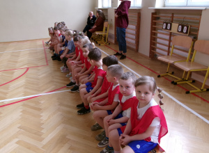 Na zdjęciu widać dzieci znajdujące się w sali gimnastycznej, ubrane w jednakowe stroje sportowe, siedzące na ławce.