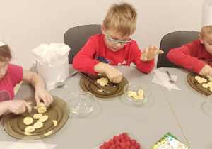 Dzieci siedzą przy stoliku i kroją banany na plasterki.