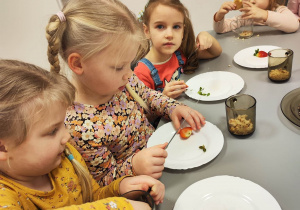 Dziewczynki kruszą ciastka do szklanki. Obok na talerzykach leżą truskawki do dekoracji.