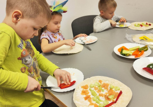 Na pierwszym planie chłopiec kroi paprykę. Obok, na talerzu, leży tortilla posmarowana sosem, na której znajdują się pokrojone warzywa.