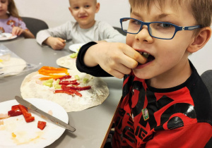 Chłopiec je kawałki warzyw. Obok, na talerzu, leży tortilla posmarowana sosem, na której znajdują się pokrojone warzywa.