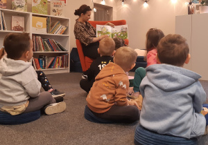 Na zdjęciu widać dzieci siedzące w bibliotece, obserwujące Panią, która czyta książkę.