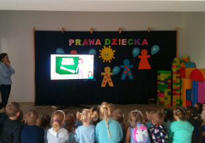 Dzieci siedzą na ławeczce na sali gimnastycznej i oglądają filmik o prawach i obowiązkach dziecka.