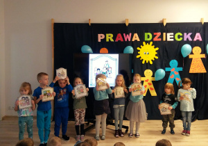 Wybrane dzieci stoją i trzymają plansze przedstawiające prawa dziecka. Pozostałe dzieci siedzą na ławeczce.