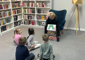 Zajęcia w bibliotece. Czwórka dzieci ogląda książki pokazywane przez Panią Bibliotekarkę. W tle znajdują się regały z książkami,