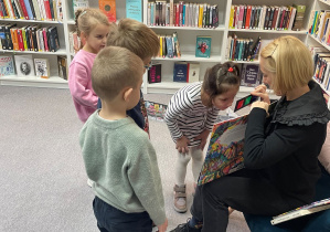 Zajęcia w bibliotece. Czwórka dzieci ogląda książki pokazywane przez Panią Bibliotekarkę. W tle znajdują się regały z książkami,