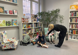 Chłopcy siedzą na dywanie, a dziewczynka w małym fotelu. Dzieci oglądają książki. Pani Bibliotekarka rozmawia z chłopcem.