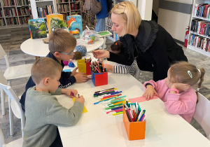 Dzieci i Pani Bibliotekarka siedzą przy stoliku, na którym znajdują się włóczka, mazaki i kredki. Dzieci ozdabiają wycięte z papieru zakładki do książek.