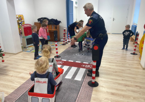 Mini miasteczko ruchu drogowego rozłożone na sali gimnastycznej. Na podłodze rozłożona jest droga, przejście dla pieszych, sygnalizacja świetlna. Dzieci poruszają się po drodze w przebraniach samochodów. Pan ze Straży Miejskiej tłumaczy dzieciom zasady bezpieczeństwa.