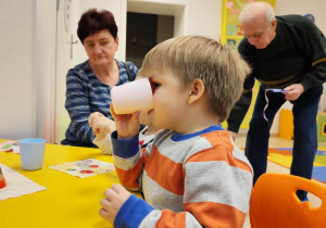 Chłopiec pije z kubka. Obok niego siedzi babcia. Na dalszym planie znajduje się dziadek.